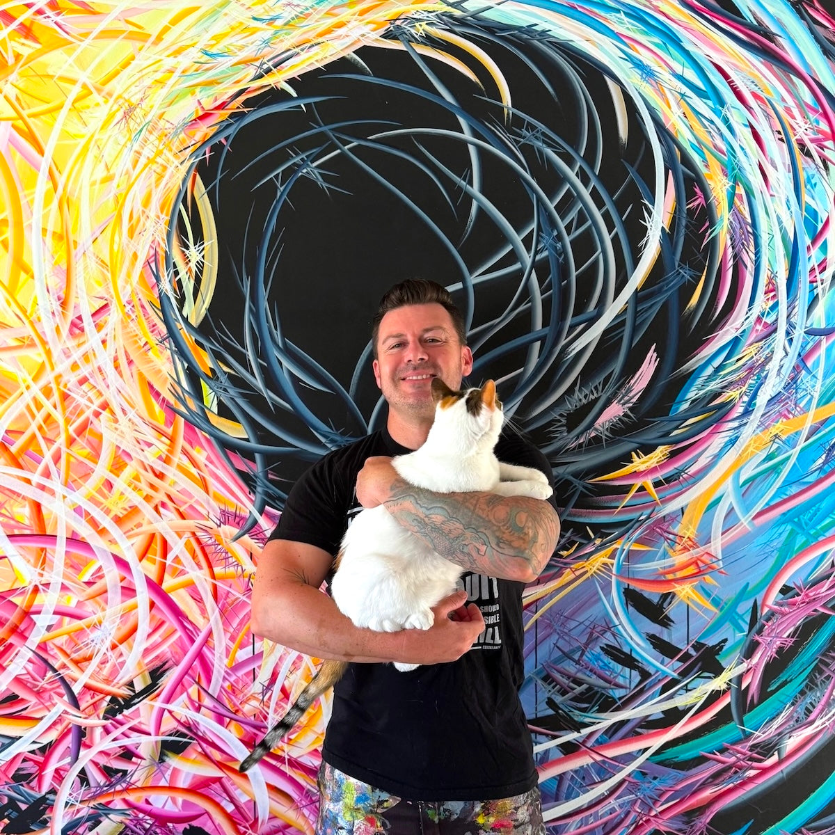 Michael Carini holding his white cat Braska in front of a colorful abstract painting